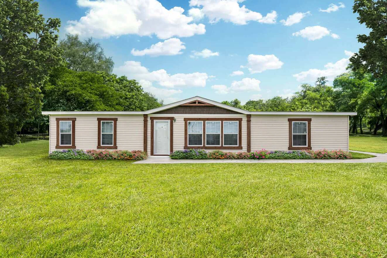 A single-story manufactured home with beige siding, brown trim, and a white front door, surrounded by green lawn, trees, and a small flower bed in front.