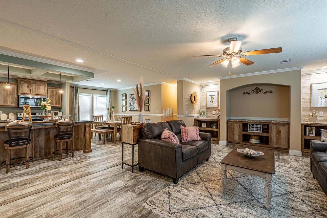 Open-concept living area with wooden floors, a kitchen with barstools, dining table, two sofas, coffee table, ceiling fan, and built-in shelves with decorations.