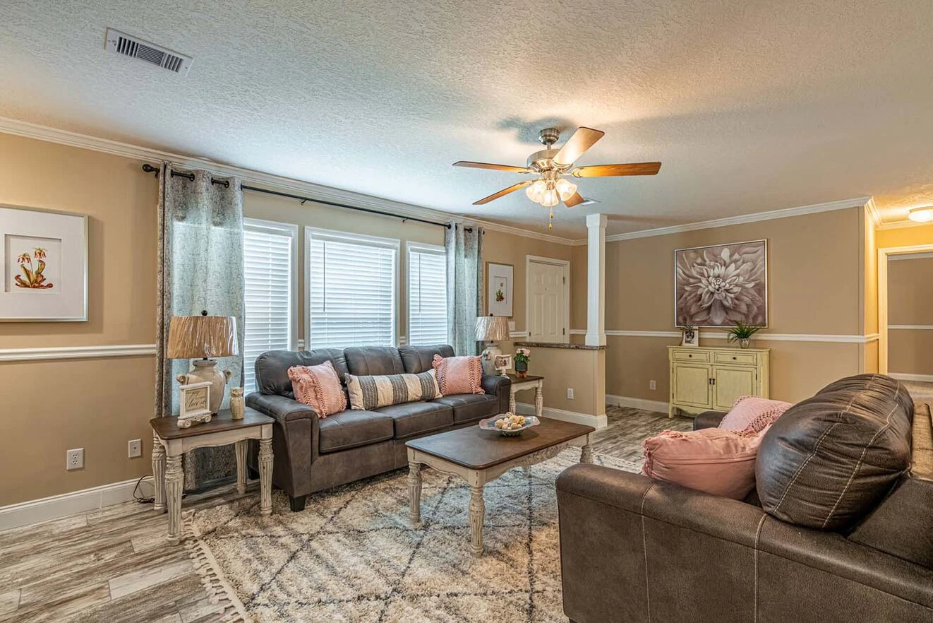 Living room with two brown sofas, a coffee table, side tables with lamps, light-colored rug, ceiling fan, large window with curtains, and wall art above a cabinet.