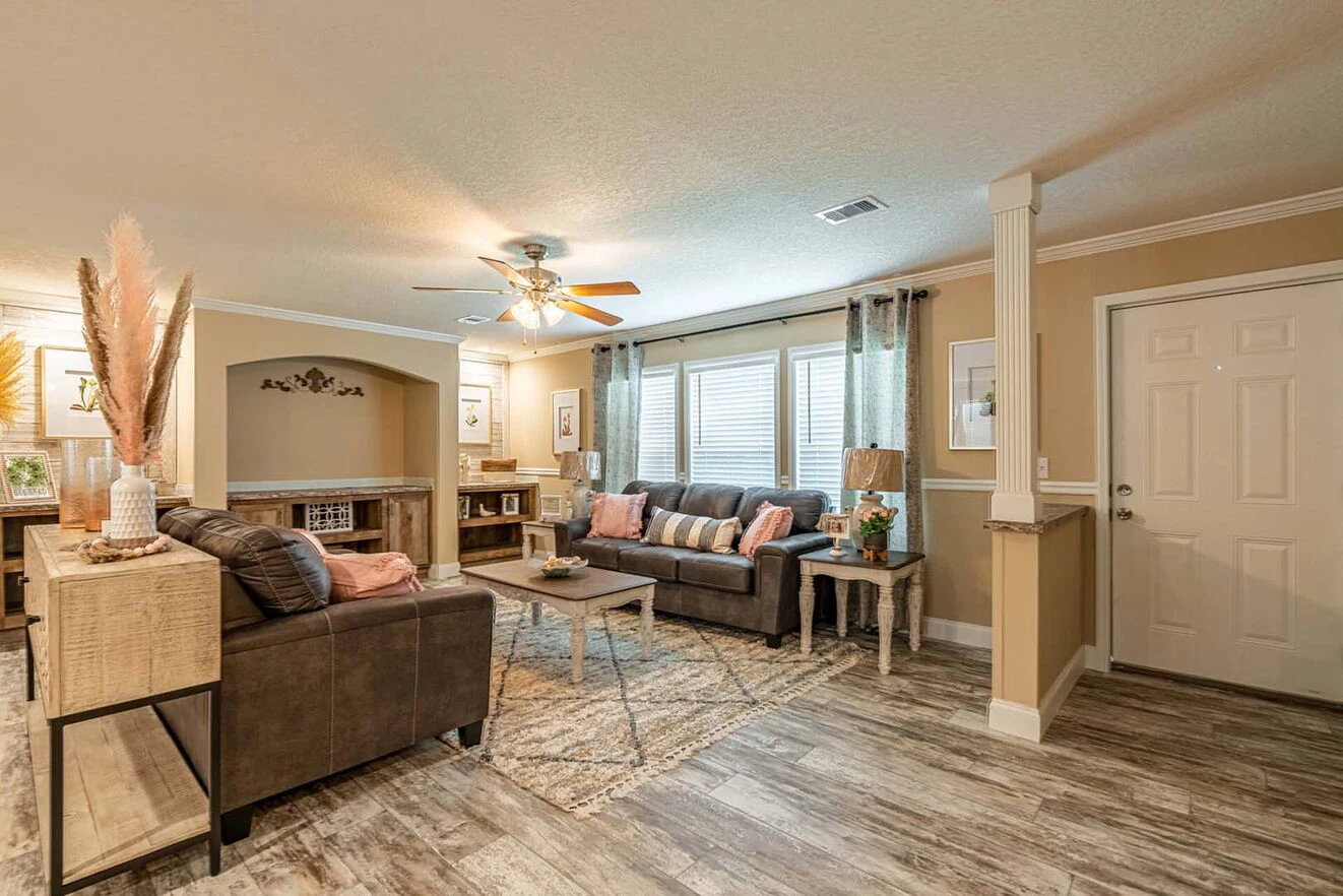 A cozy living room with two brown sofas, a coffee table, decorative plants, wall art, large windows with curtains, ceiling fan, and wood-style flooring.