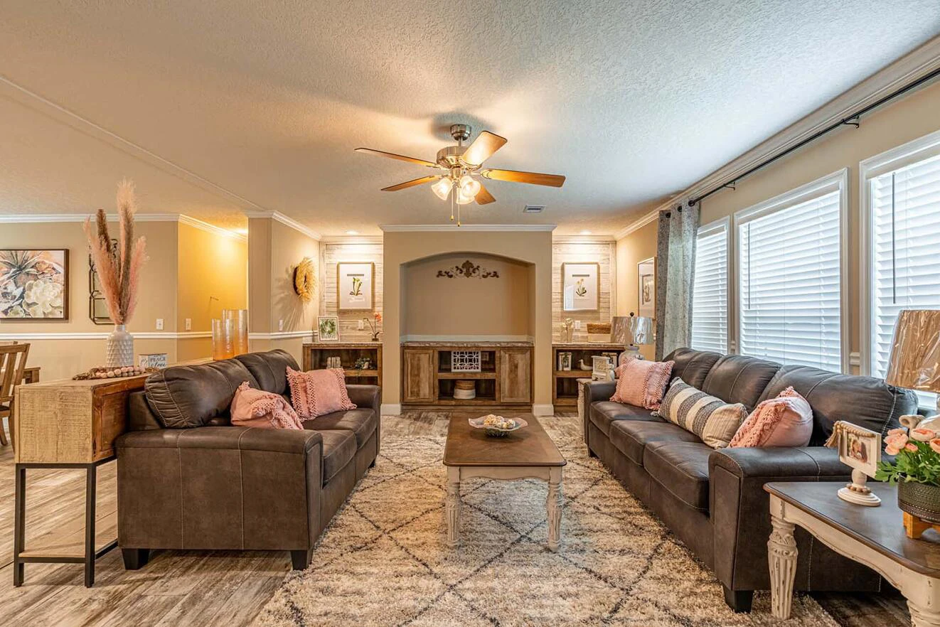 A cozy living room with two brown sofas facing a wooden coffee table, neutral decor, large windows with blinds, and a ceiling fan with lights.