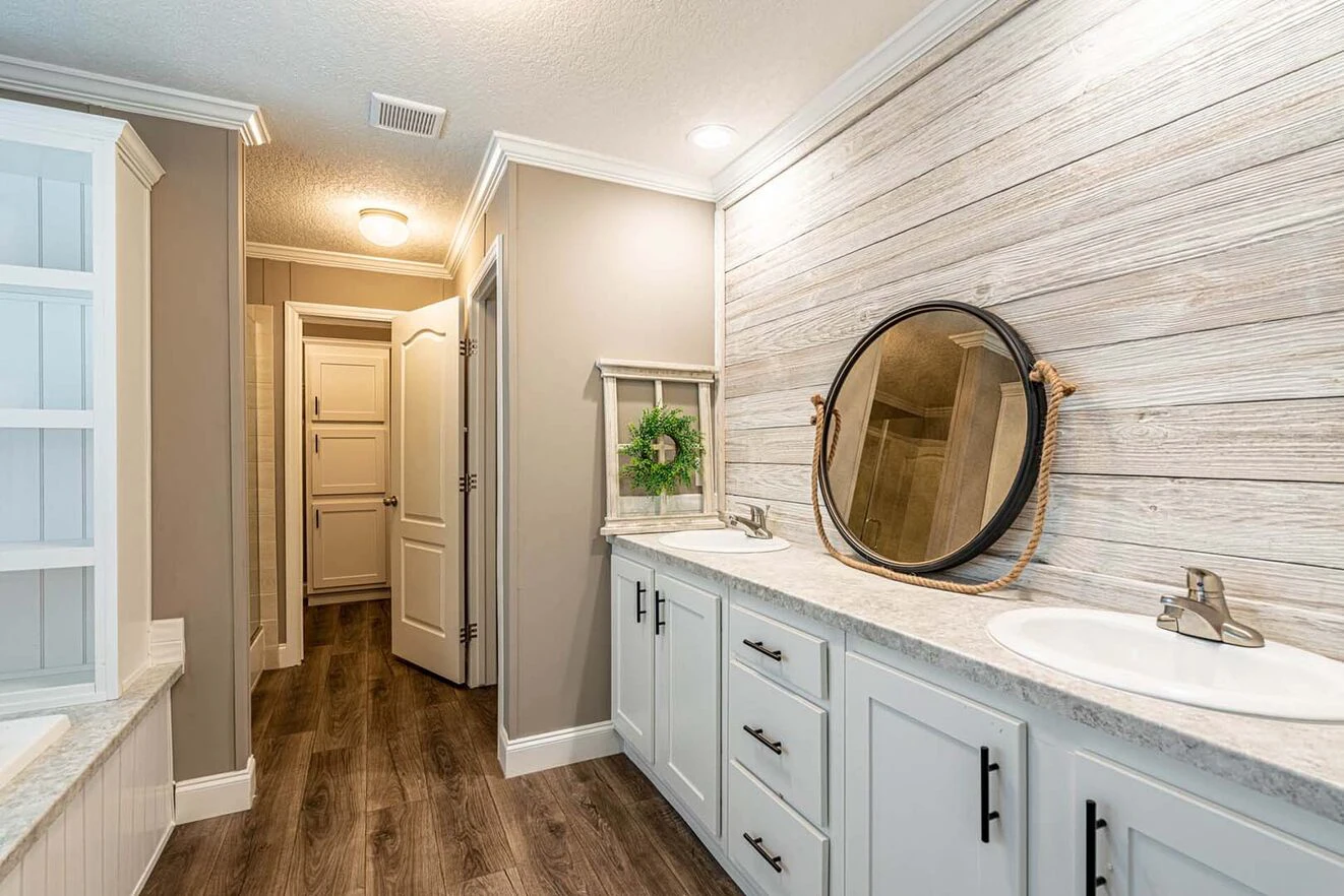 Modern bathroom with wood-look flooring, double sinks, round mirror with rope, white cabinets, and light wood accent wall; small greenery decor beside the sink.
