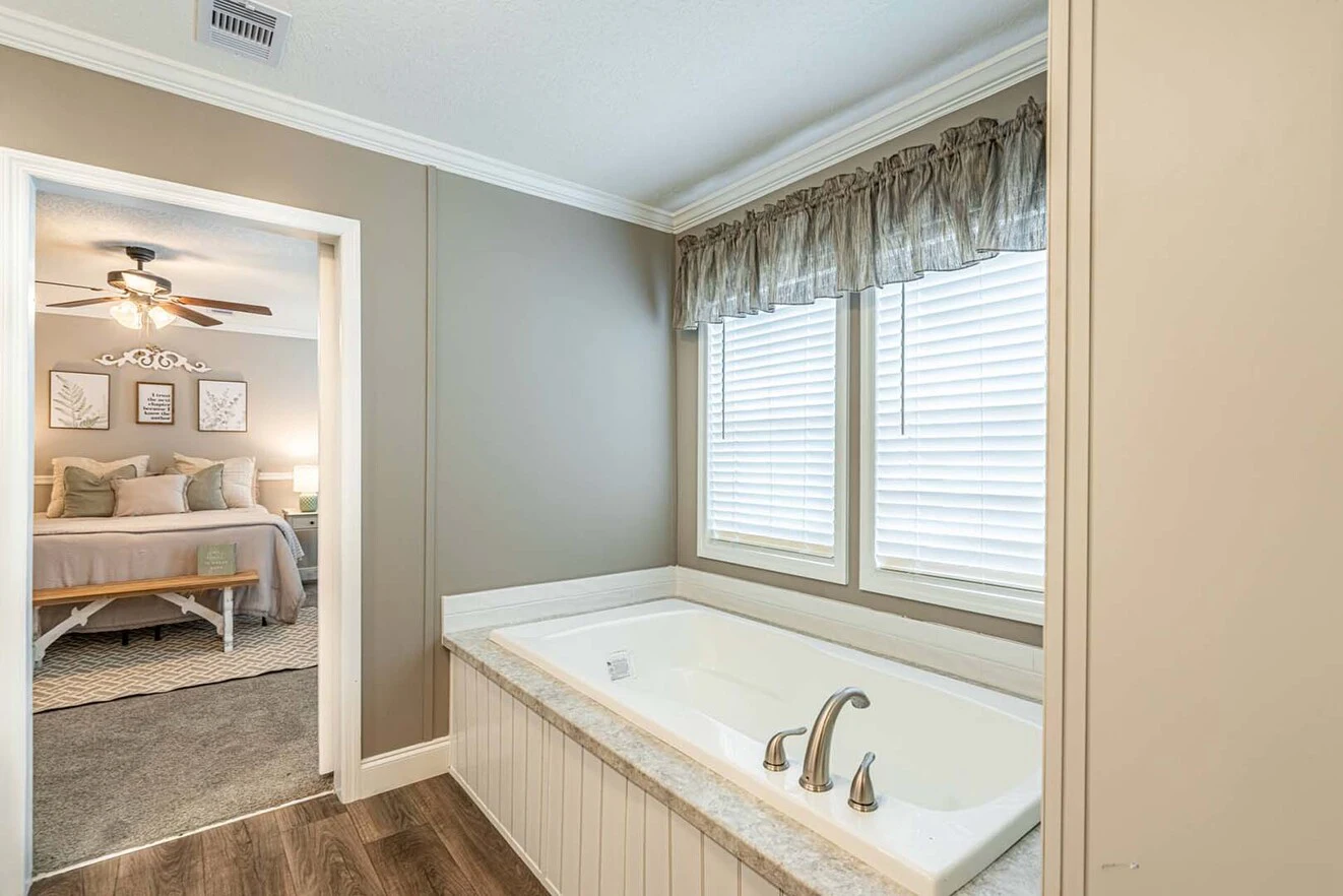 A bathroom with a built-in bathtub beneath two windows, connected to a bedroom with a ceiling fan and bed visible through an open doorway.