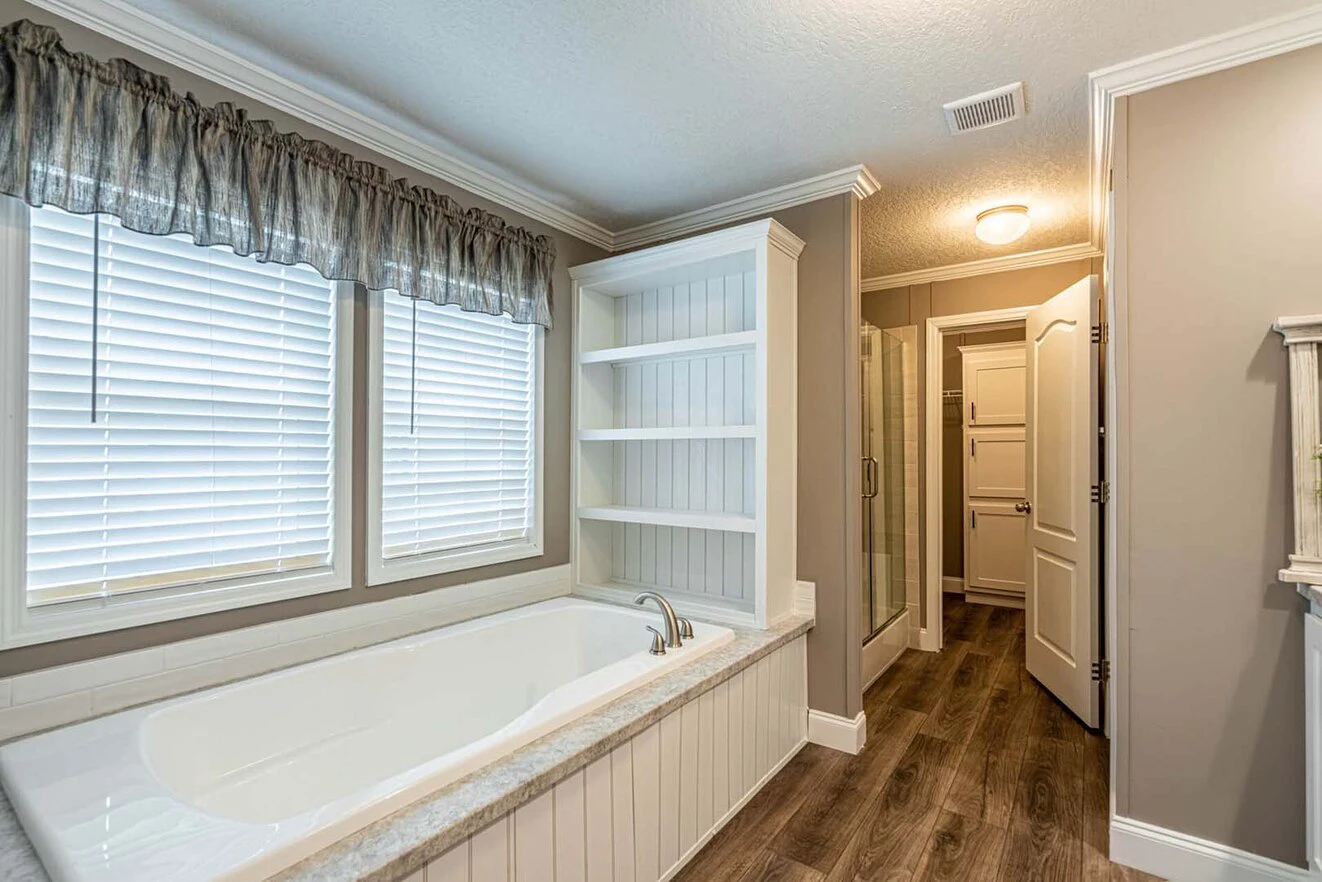 A bathroom with a built-in bathtub under two windows, open shelving, wood-look flooring, and a glass-enclosed shower in the background.