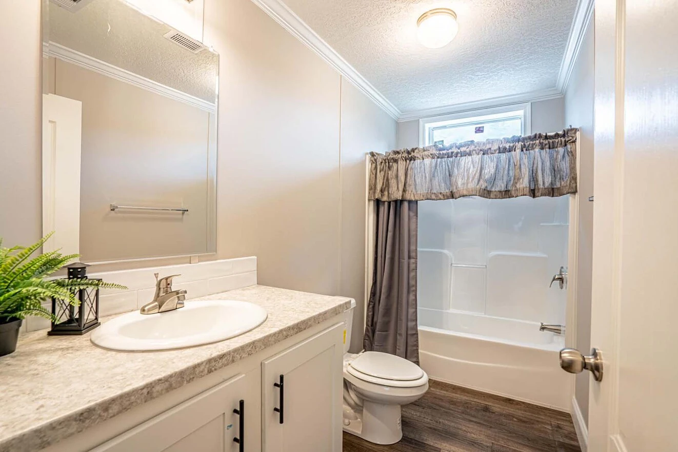 Modern bathroom with a single sink vanity, white countertop, toilet, bathtub with shower curtain, wood-look flooring, and a small window above the tub.