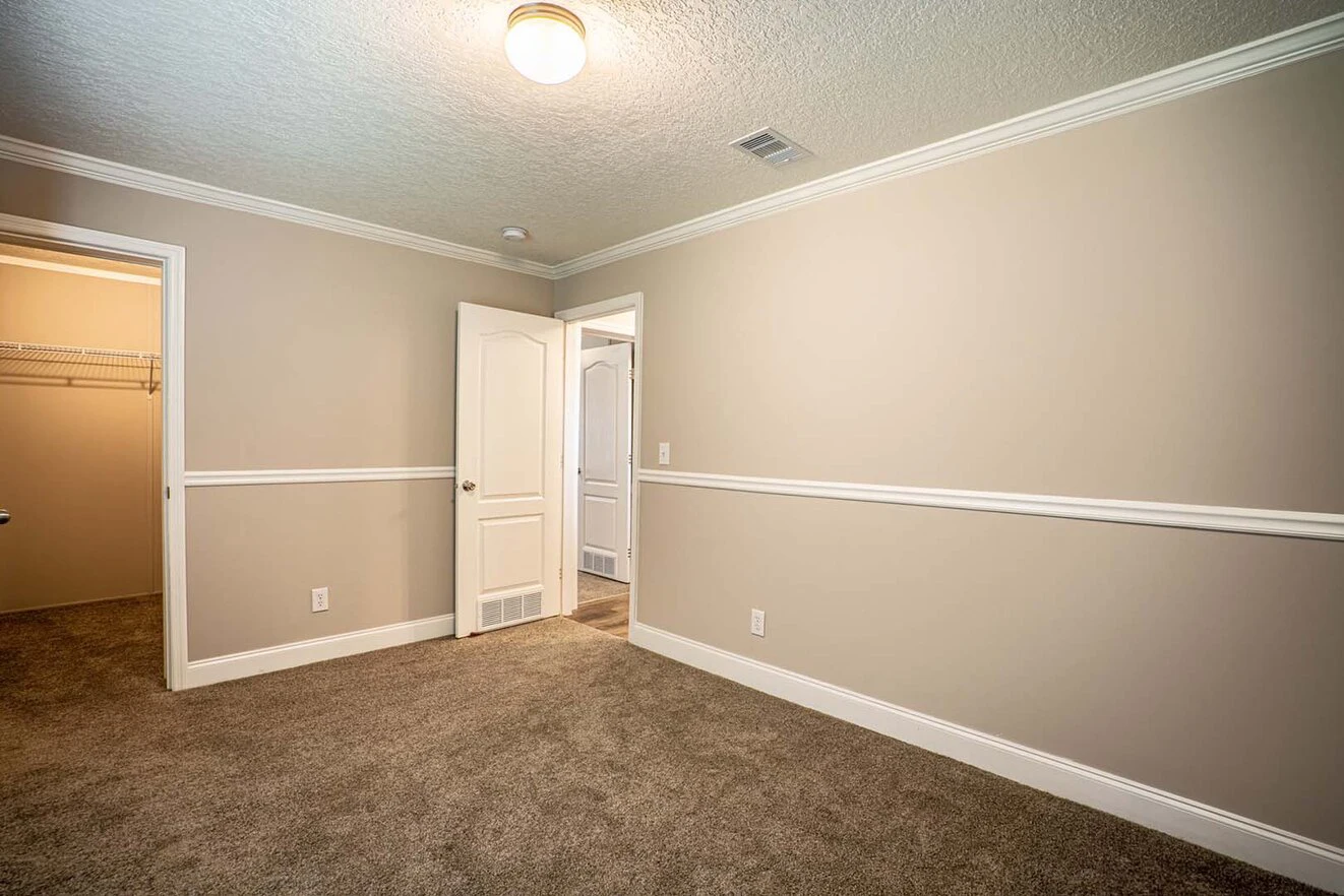 Empty beige room with carpet flooring, white trim, walk-in closet to the left, and an open door leading to a hallway. Ceiling light is on.