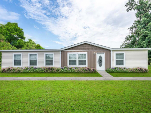 Single-story manufactured home with a gable roof, white siding, central front door, symmetrical windows, and a flower bed along the front, set on a grassy lawn.