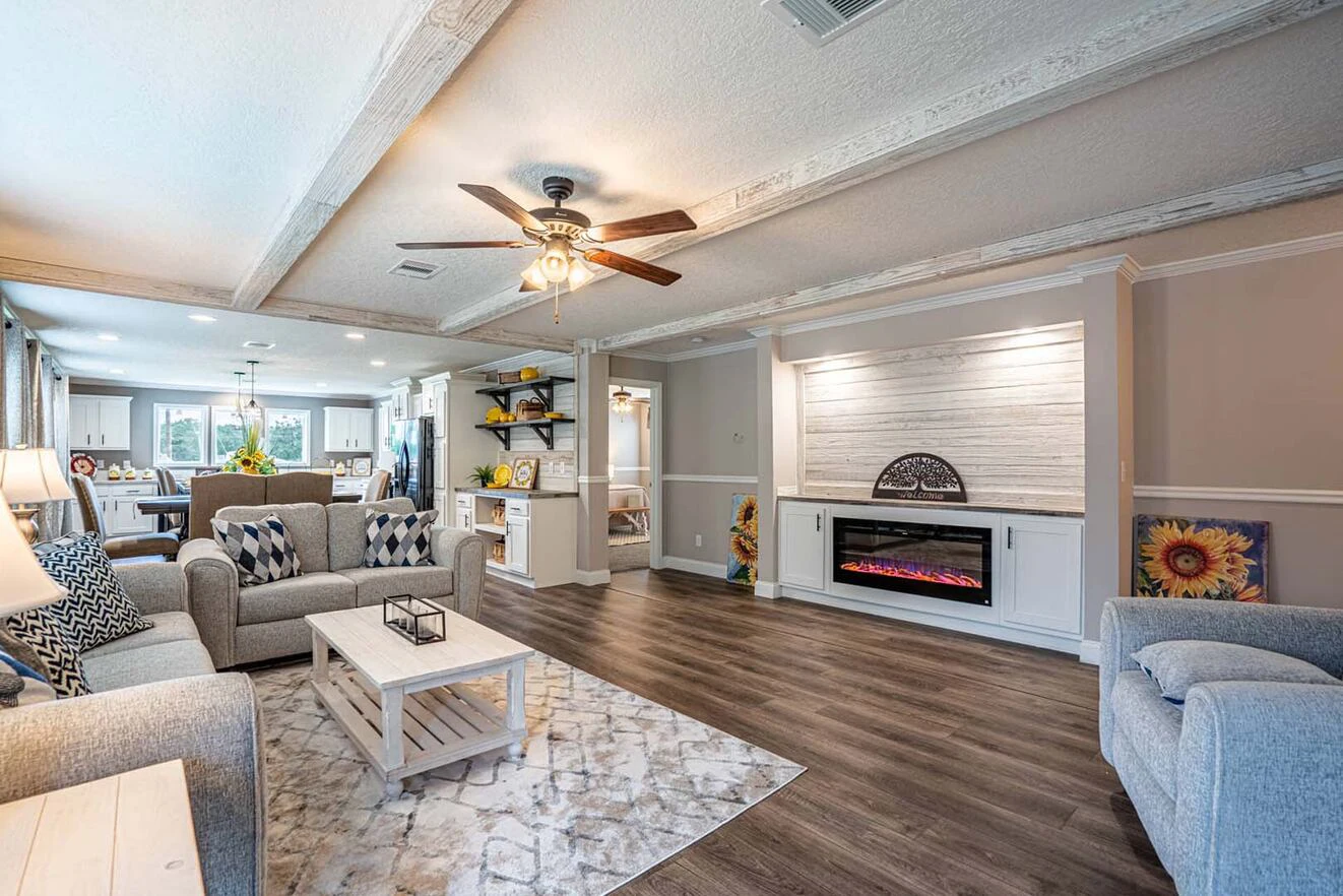 A spacious living room with gray sofas, a ceiling fan, wood flooring, a modern electric fireplace, and an open layout leading to the dining area and kitchen.