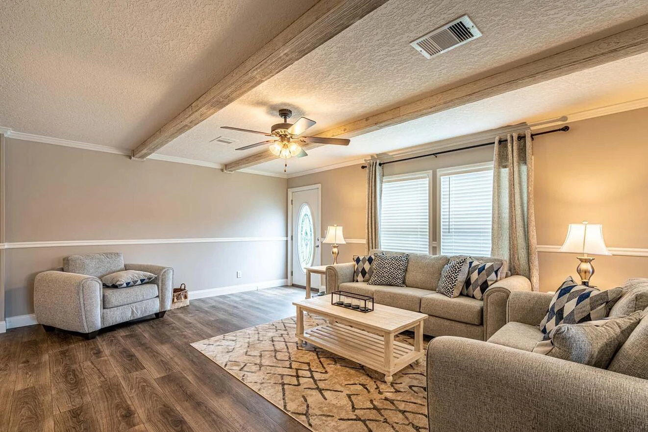 A living room with beige walls, wood ceiling beams, two sofas, an armchair, coffee table, lamps, and large windows with curtains.