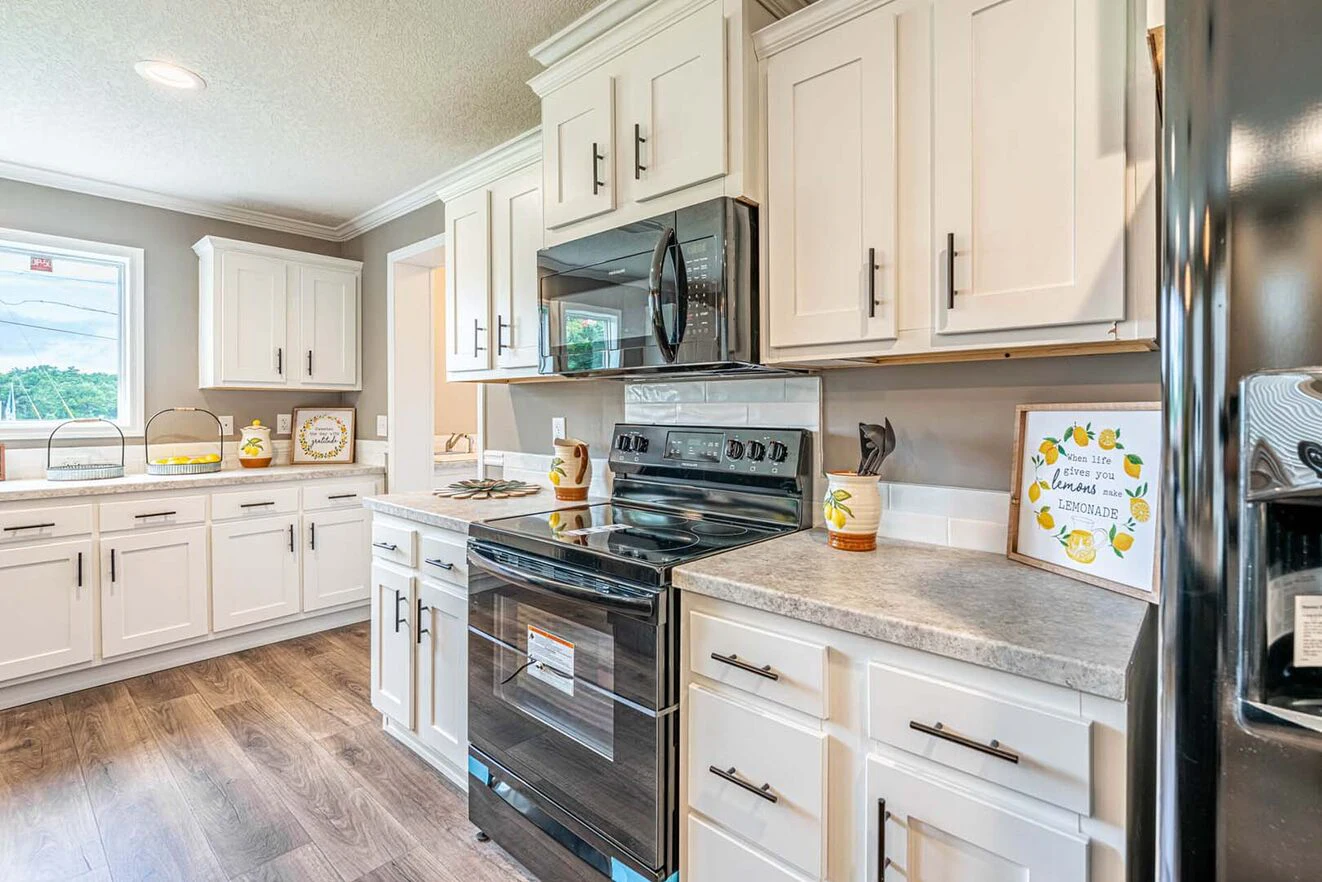Modern kitchen with white cabinets, black appliances, gray countertops, wood flooring, and a window providing natural light. Decorative items and small appliances are on the counters.