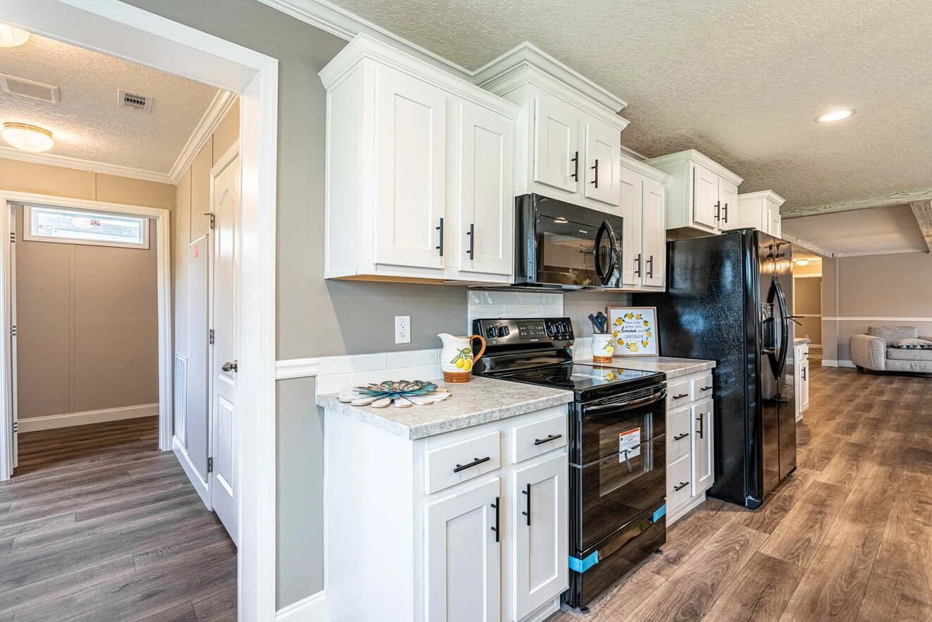 Modern kitchen with white cabinets, black appliances, marble countertops, and wood-style flooring; hallway and living area visible in the background.