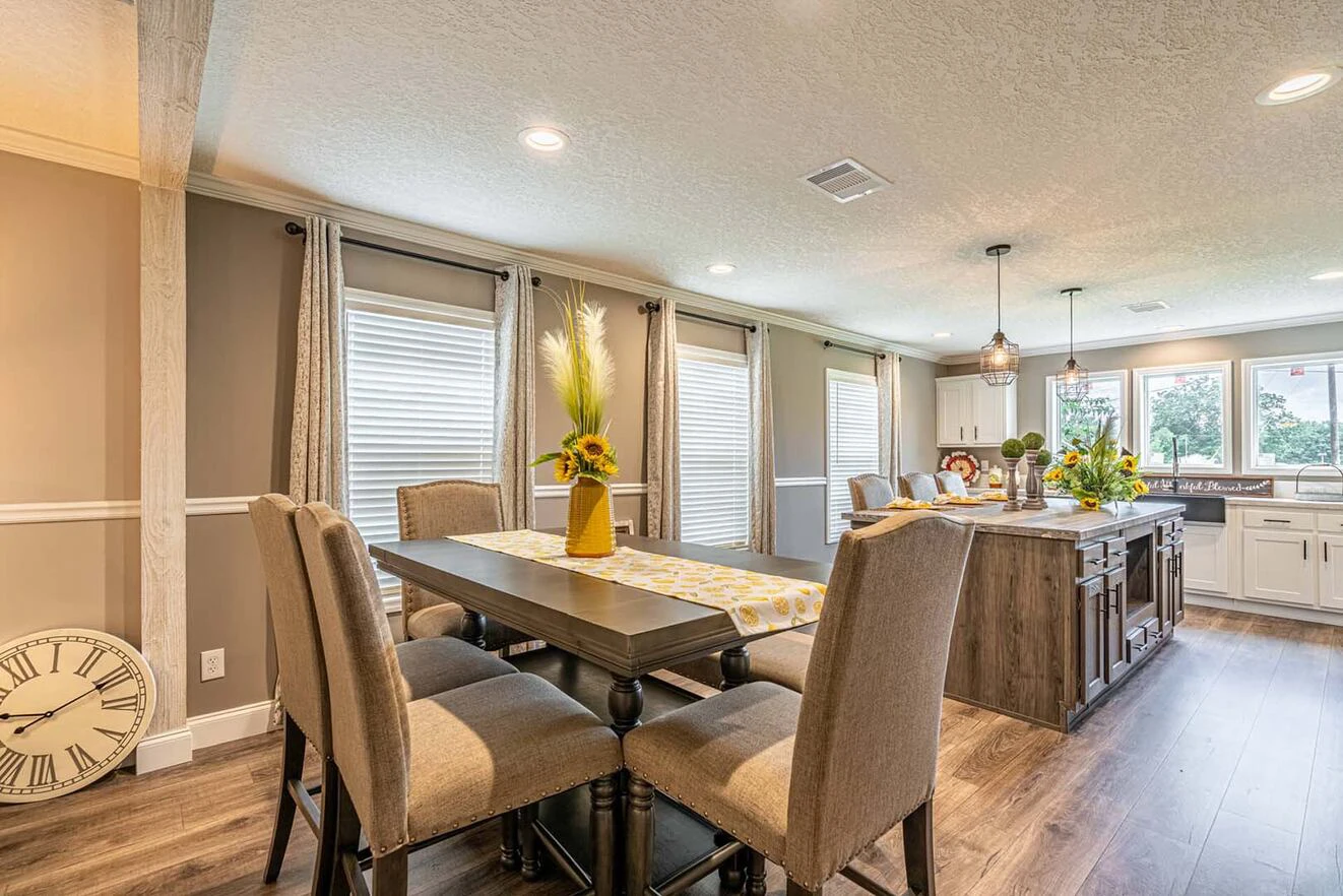 Modern dining area with upholstered chairs and a wooden table, decorated with sunflowers, next to a bright open kitchen with an island and large windows.