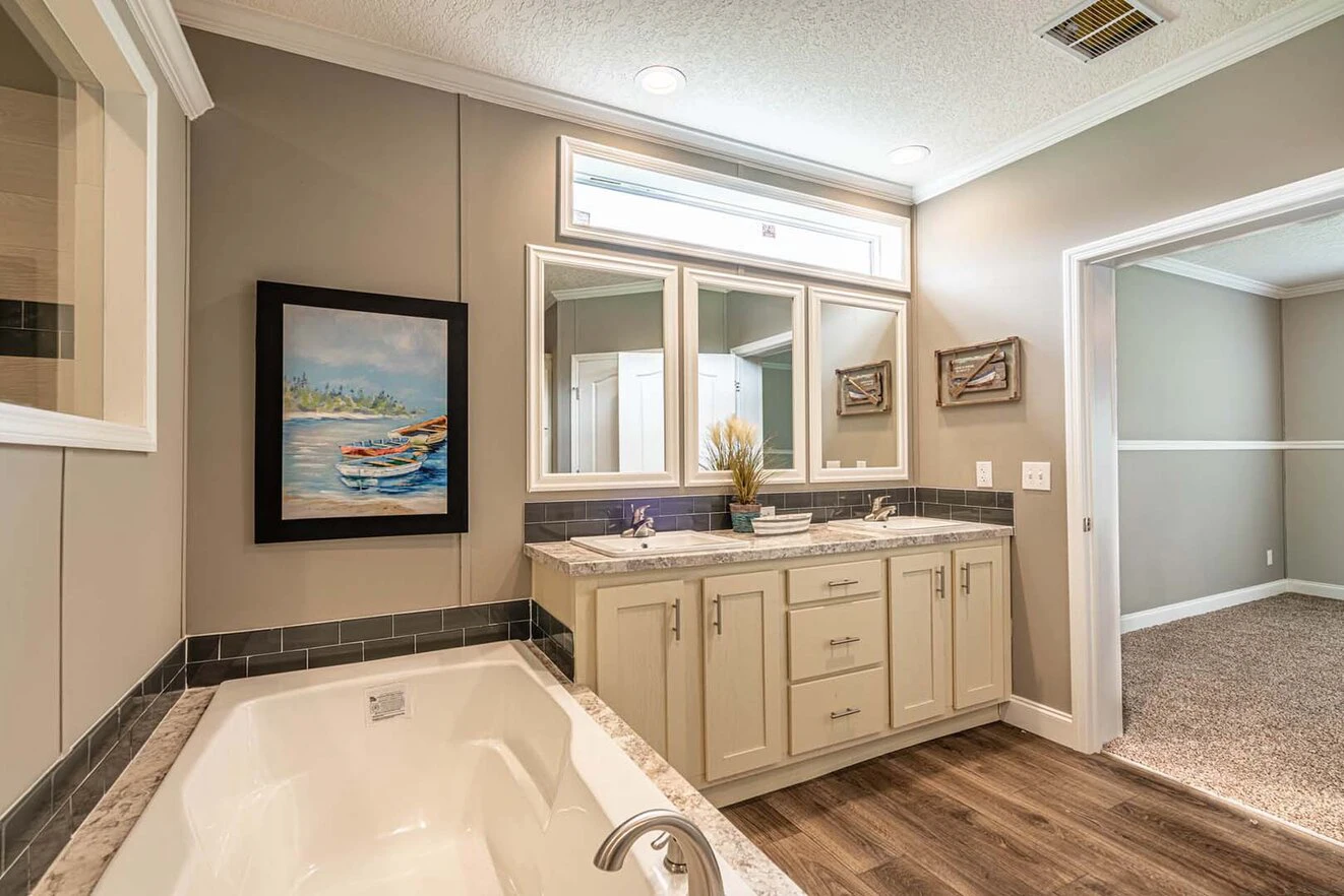 Modern bathroom with a soaking tub, double vanity with mirrors, neutral walls, wood flooring, and a painting of a boat on the wall. An open doorway leads to a carpeted room.