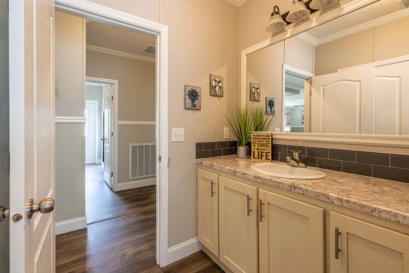 A bathroom with a single sink vanity, large mirror, beige cabinets, gray tile backsplash, and a view into a hallway with wood flooring.