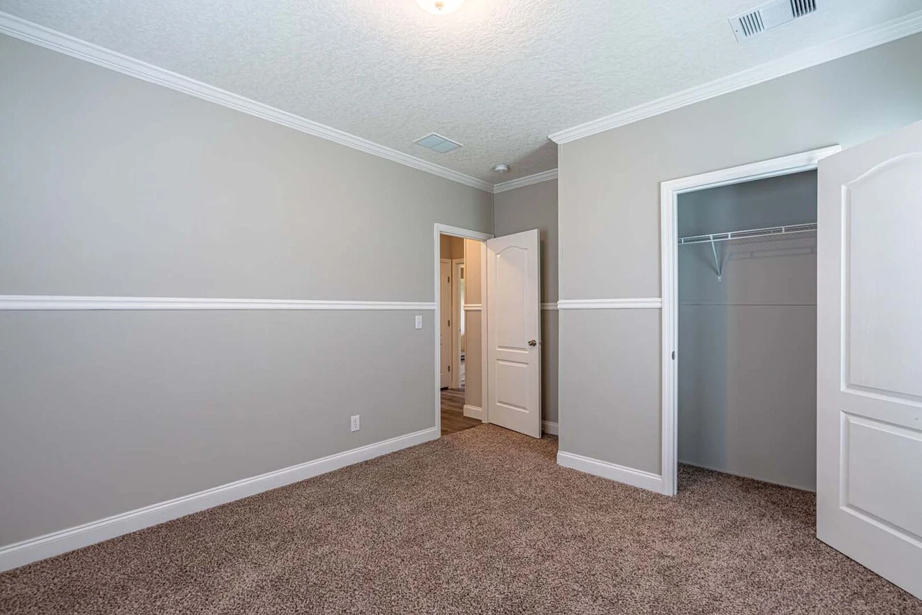 Empty bedroom with beige carpet, light gray walls, white trim, an open closet door, and an open door leading to a hallway.