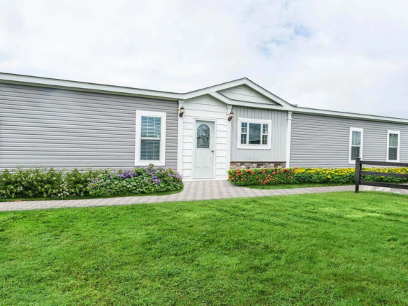 Single-story, light gray manufactured home with white trim, centered front door, and flower beds along a paved walkway, surrounded by green lawn.