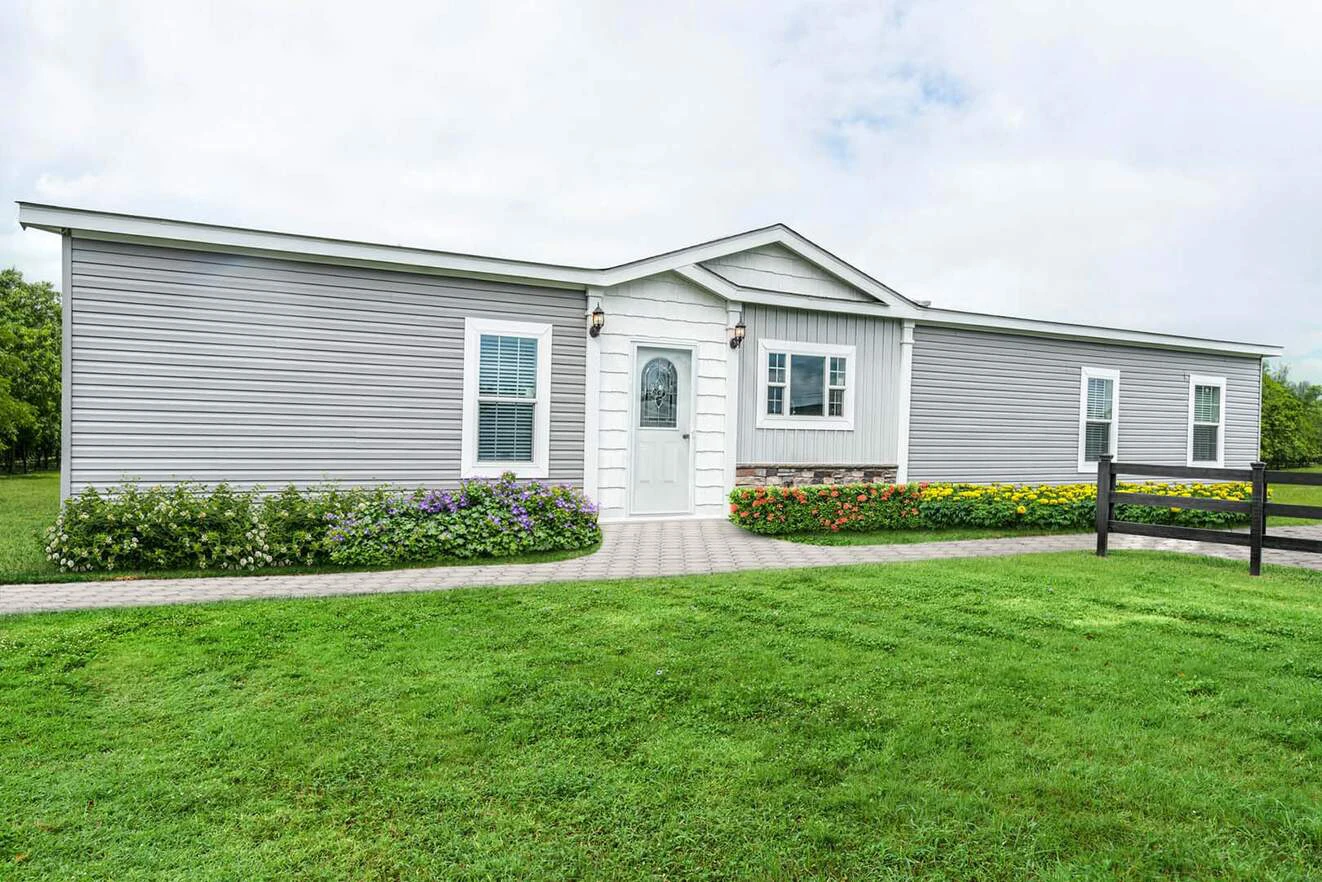 Single-story, light gray manufactured home with white trim, centered front door, and flower beds along a paved walkway, surrounded by green lawn.