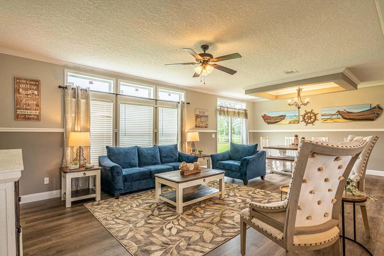Living room with blue sofas, armchairs, coffee table, and rug; dining area in the background with nautical decor and large windows letting in natural light.