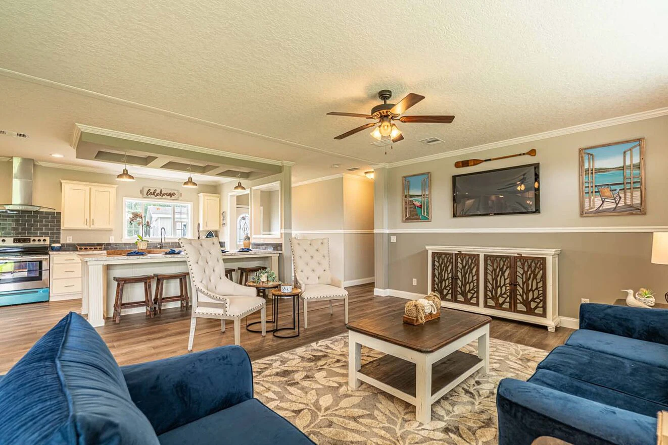 Spacious living room with blue sofas, a wooden coffee table, a TV mounted on the wall, and an open kitchen in the background. Ceiling fan and light fixture overhead.