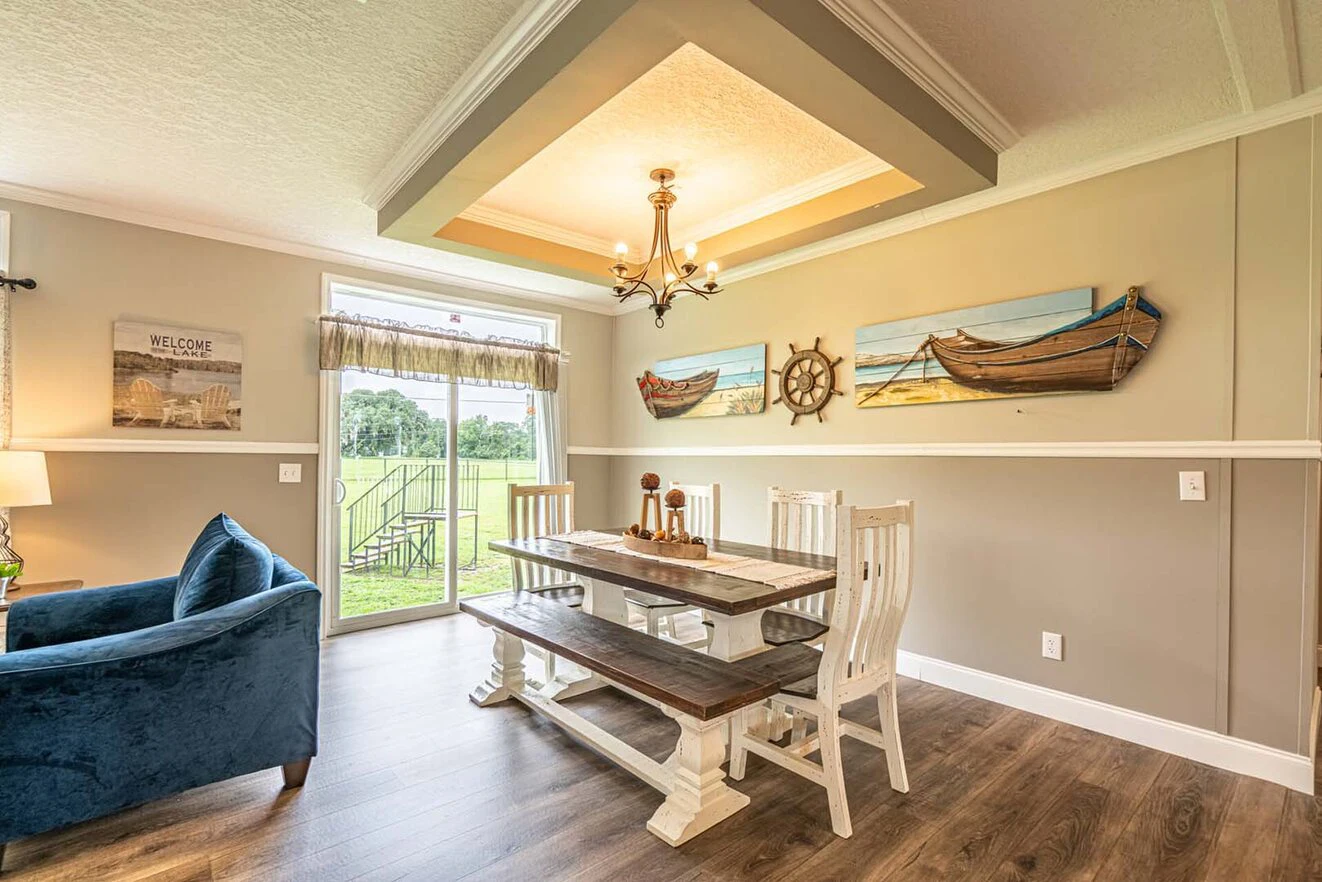 A dining area with a wooden table, bench seating, nautical-themed wall art, chandelier, blue sofa, and large window overlooking a grassy yard.
