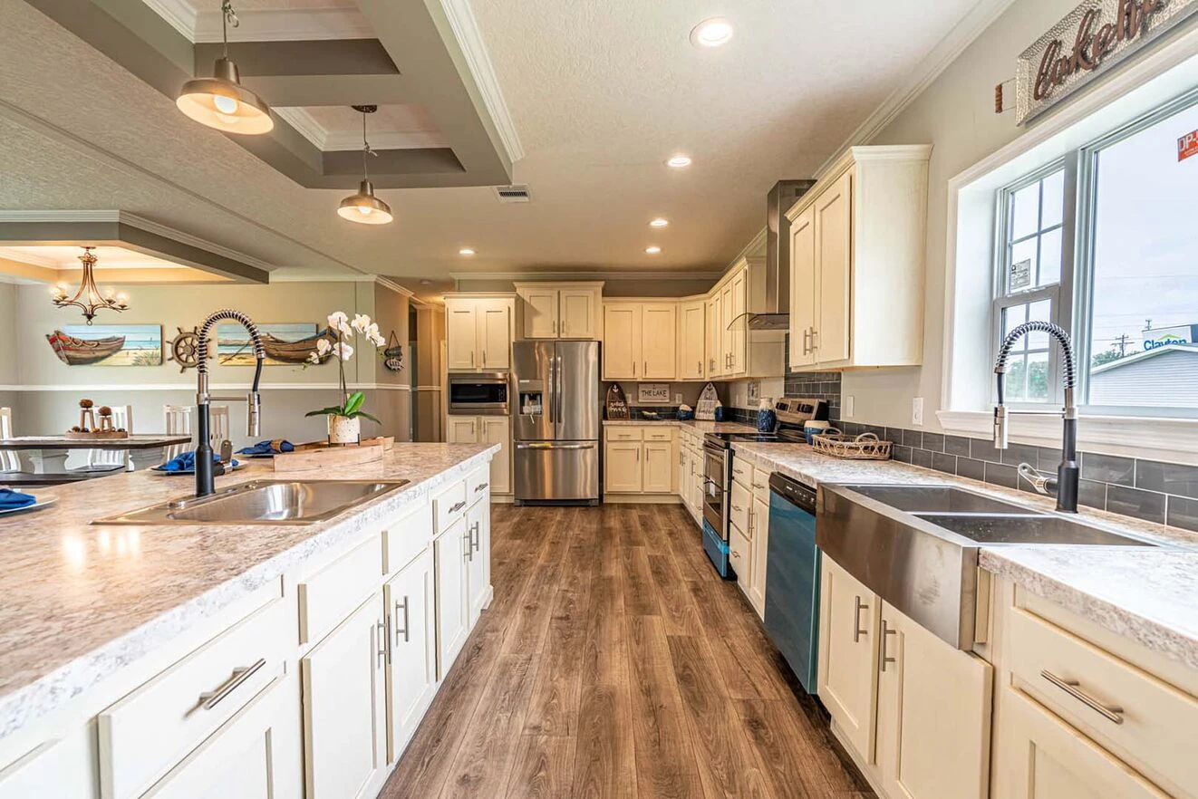 Modern kitchen with white cabinets, stainless steel appliances, double sinks, marble countertops, and wood flooring, viewed from the center island toward a window and dining area.