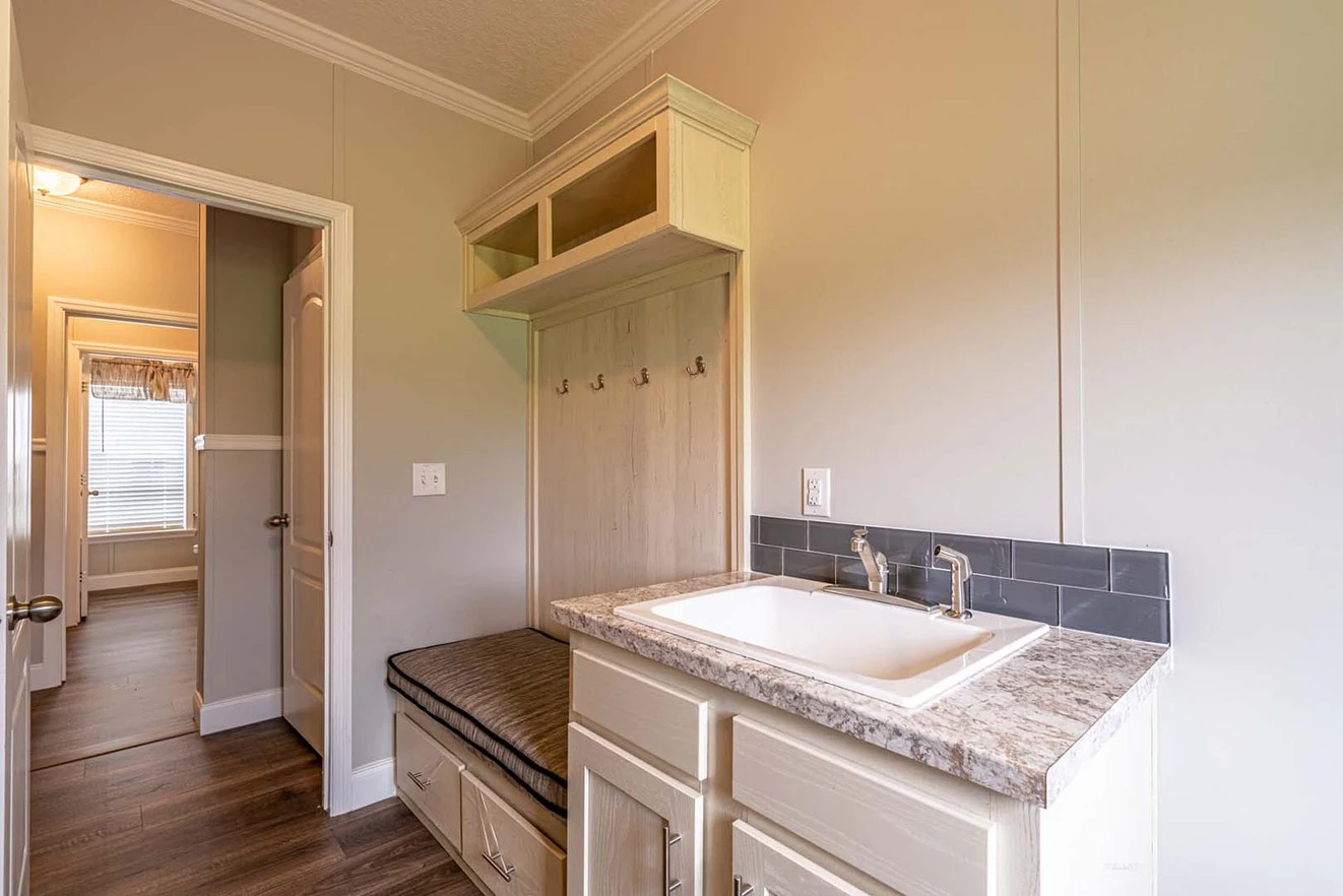 A mudroom with a cushioned bench, hooks for hanging items, overhead storage, and a sink with a granite countertop and backsplash.