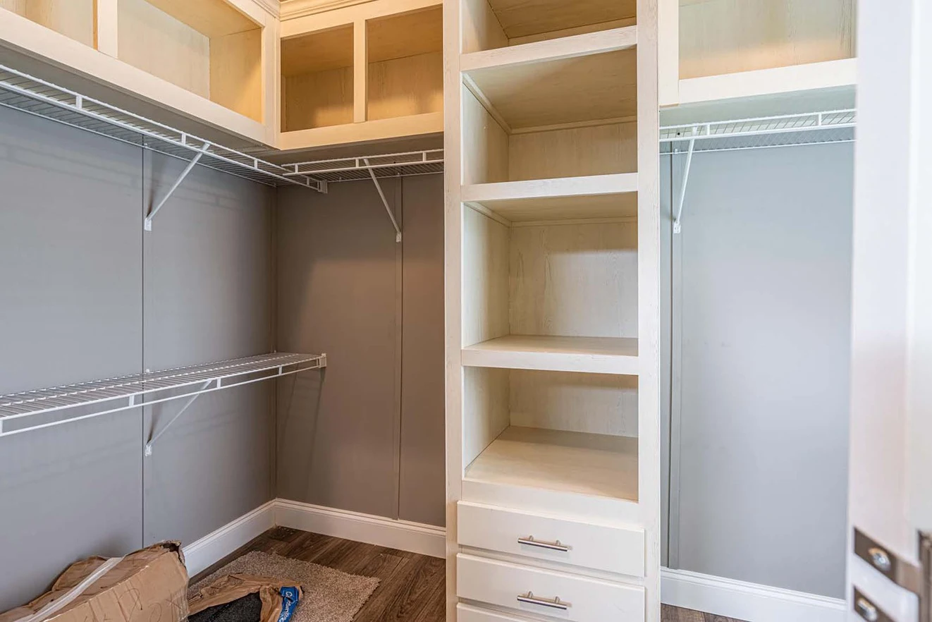 Empty walk-in closet with built-in wooden shelves, drawers, and open cabinets, plus white wire hanging racks against light gray walls.