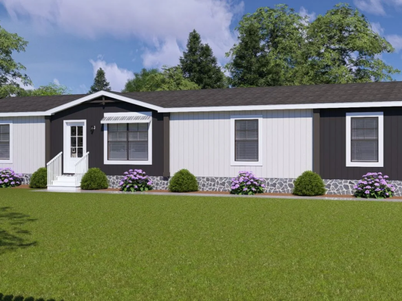 A single-story manufactured home with white and dark trim, front steps, shrubs, purple flowers, and a large green lawn. Trees and a white fence are in the background.