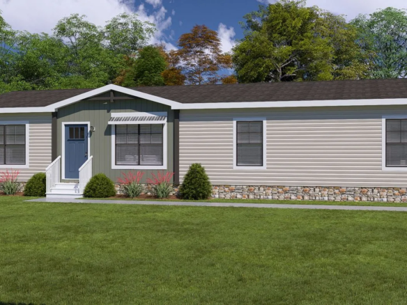 Single-story manufactured home with beige siding, stone trim, white-framed windows, and small shrubs in front yard, set against a backdrop of trees and blue sky.