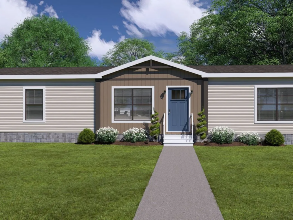 A single-story manufactured home with beige siding, a gray front door, and a small landscaped garden, set against a grassy yard and clear sky.