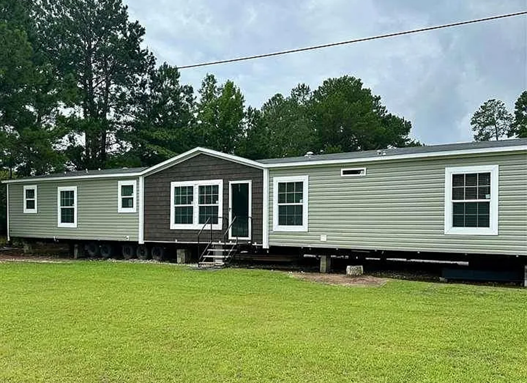A manufactured home sits on blocks and wheels in a grassy yard, surrounded by trees, with stairs leading to the front door.