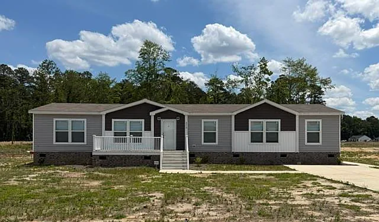 A single-story manufactured home with white siding, dark trim, a front porch, and steps, set on a sparse grassy lot under a partly cloudy sky.