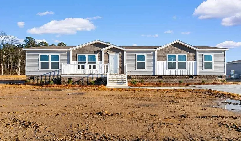 A newly constructed manufactured home with gray siding, white trim, a small porch, and stairs, set on a cleared lot under a partly cloudy sky.