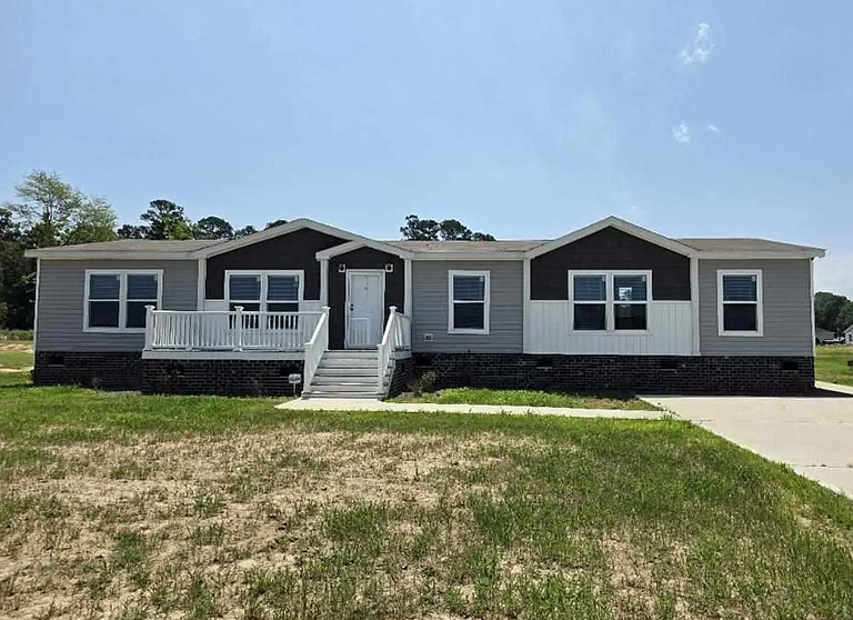 Single-story modular home with dark siding, white trim, a front porch with steps, and a concrete driveway on a partially grassy lot.