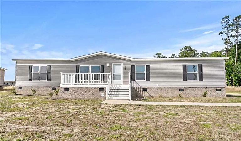 A single-story manufactured home with gray siding, white trim, front porch, and multiple windows, set on a grassy lot under a blue sky.