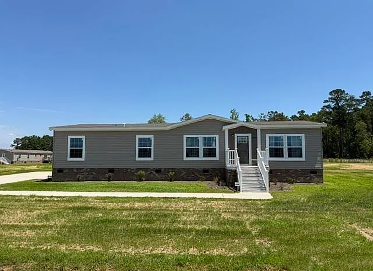 Single-story manufactured home with gray siding, white trim, and a front porch with steps, set on a grassy lawn under a clear blue sky.