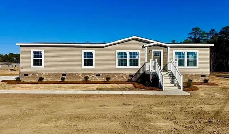 A beige manufactured home with white trim, brick foundation, multiple windows, and stairs leading to the front door, set on a dirt lot under a clear blue sky.