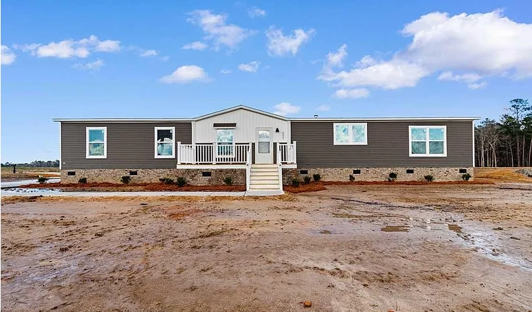 A modern manufactured home with a gray exterior, white trim, and front porch sits on a cleared lot under a partly cloudy sky.