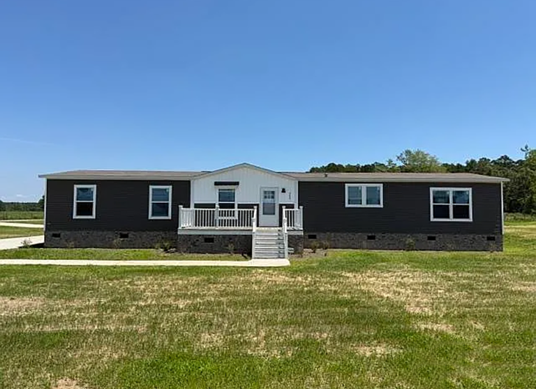 A single-story manufactured home with dark siding, white trim, and a small front porch sits on a grassy lot under a clear blue sky.