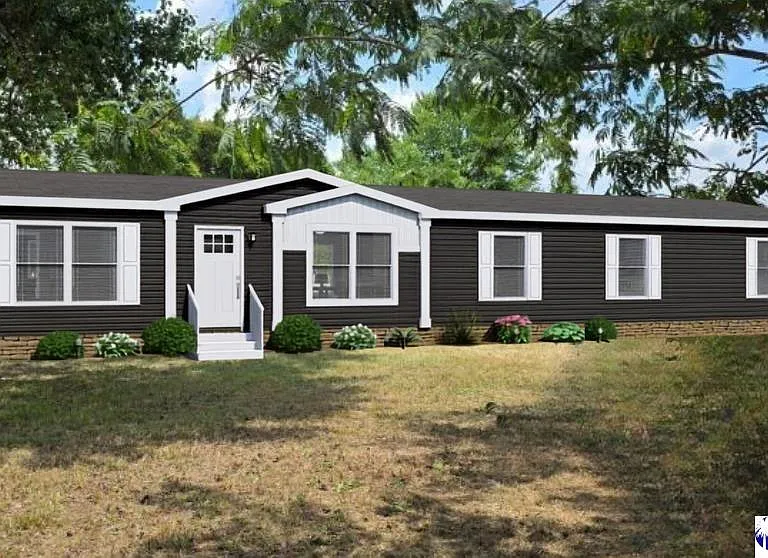 A single-story, dark brown manufactured home with white trim, multiple windows, a small front porch, and minimal landscaping on a grassy lot with surrounding trees.