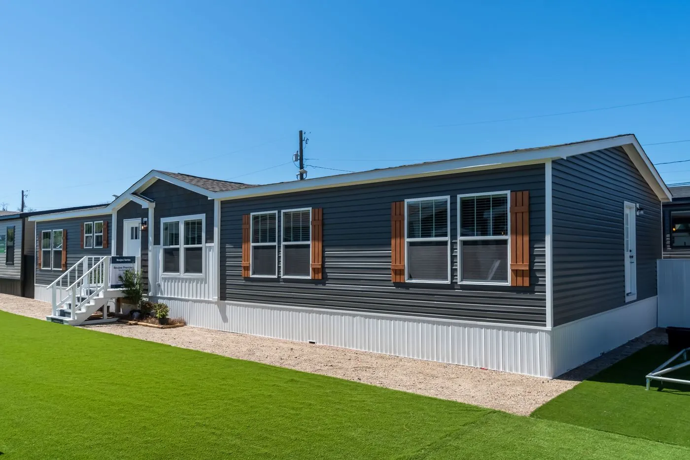 A modern manufactured home with dark gray siding, white trim, wood shutters, and artificial grass lawn under a clear blue sky.