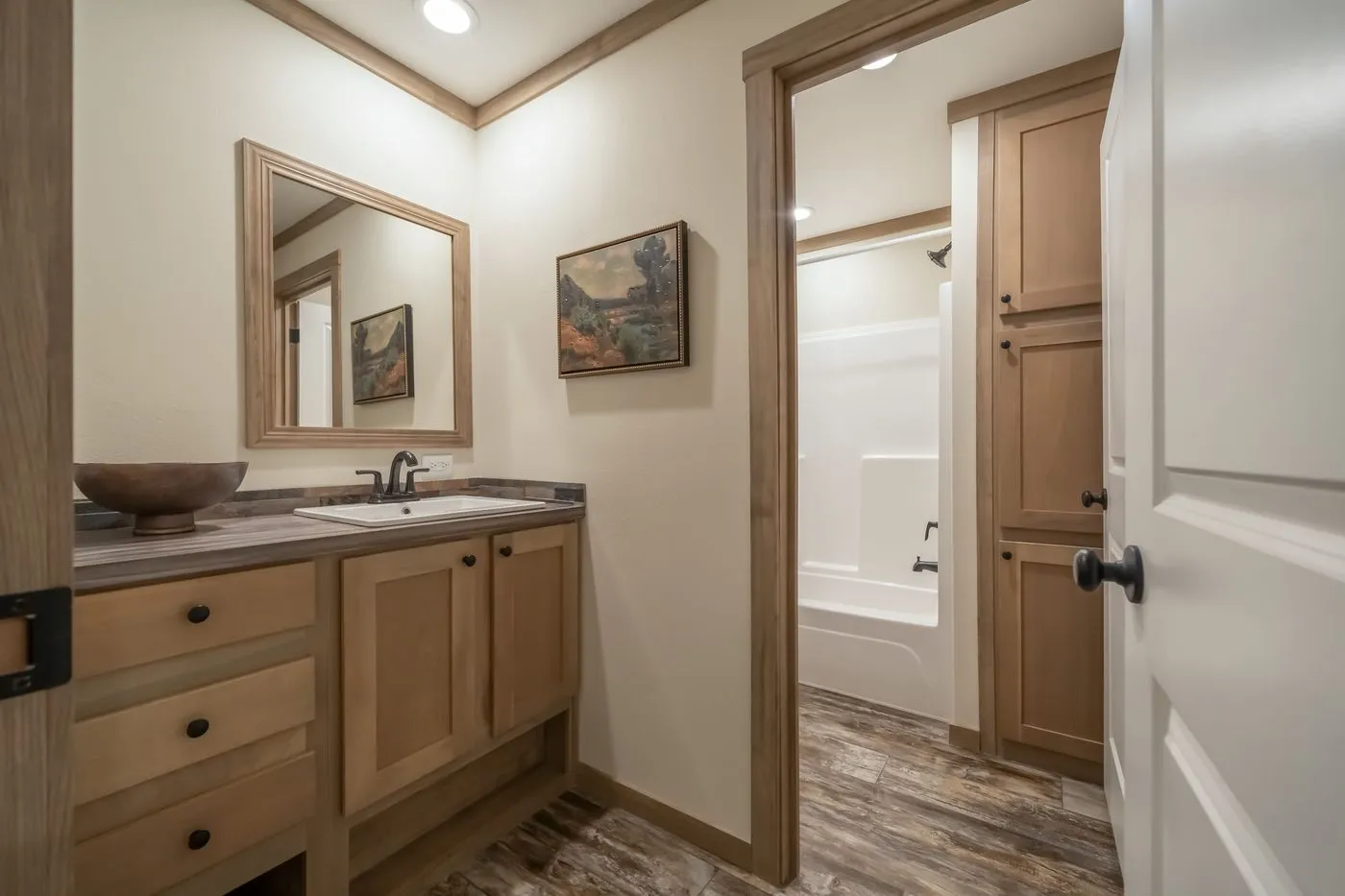 Bathroom with wood cabinets, a vanity with a mirror, countertop with a bowl, and a separate room with a white bathtub and shower. Neutral tones and tile flooring.