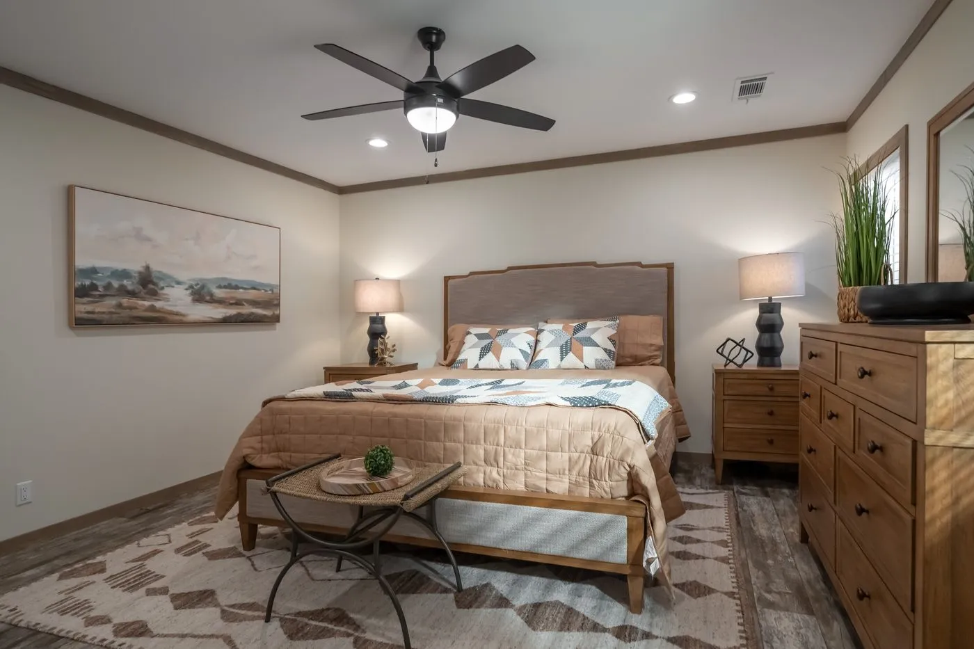 A neatly made bed with geometric bedding, two nightstands with lamps, a ceiling fan, dresser with plants, and landscape artwork in a modern bedroom.