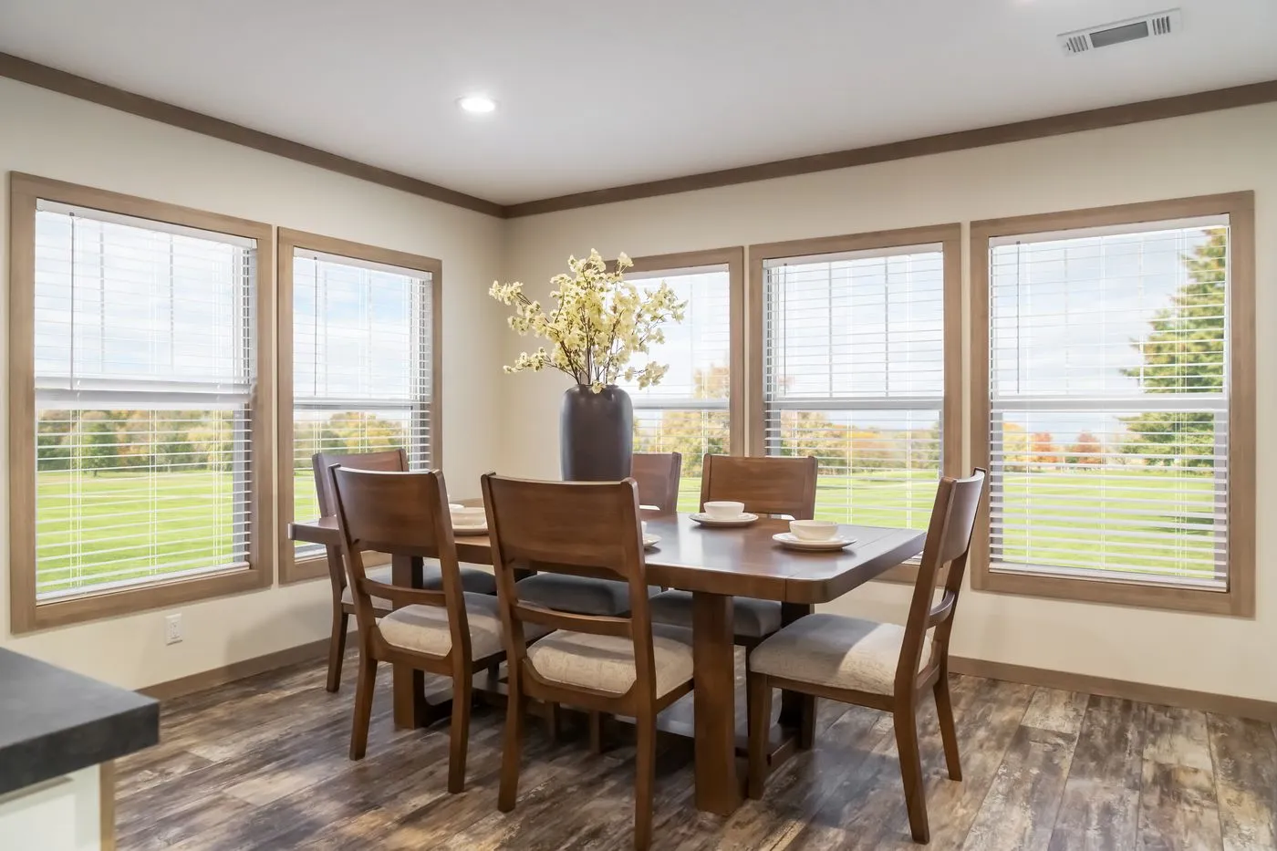 A dining room with a dark wooden table, six chairs, a vase with flowers, and large windows showing a green outdoor view.