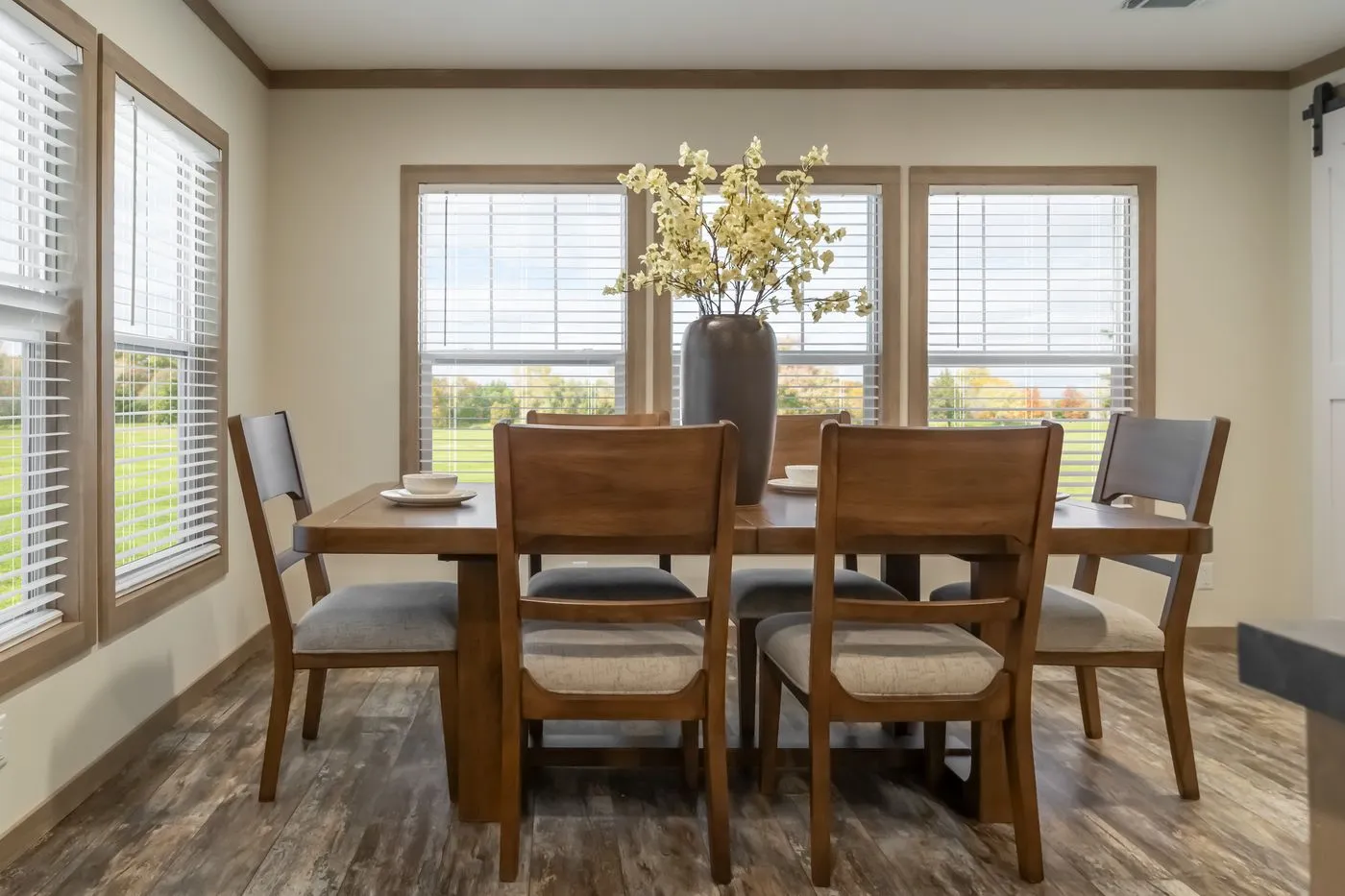 A wooden dining table with six chairs is set with plates and bowls, featuring a large vase with white flowers as a centerpiece, in a bright room with large windows.