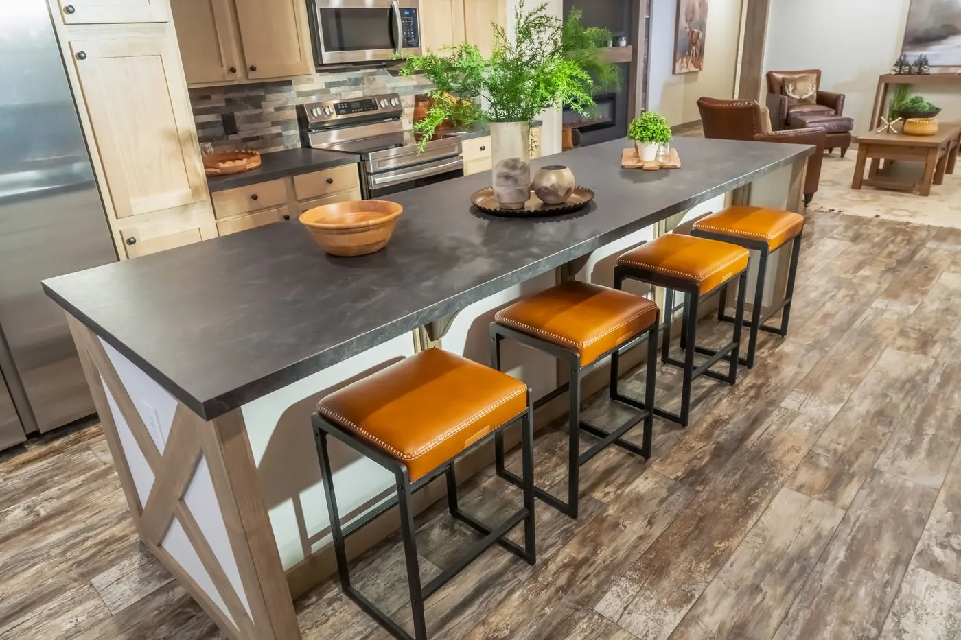 A modern kitchen features a long island with a dark countertop, four orange cushioned stools, wooden cabinetry, and stainless steel appliances.