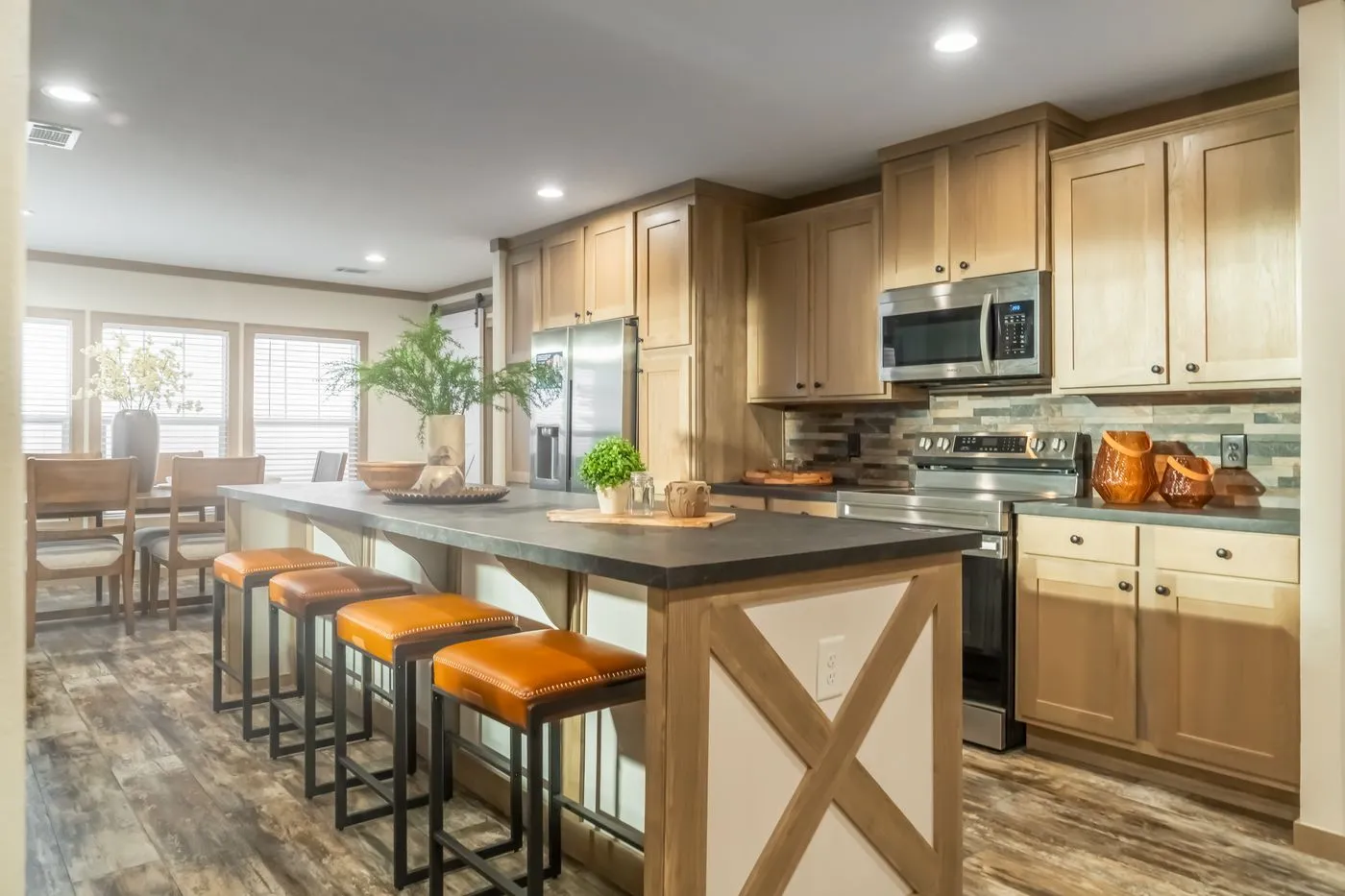 Modern kitchen with wood cabinets, stainless steel appliances, a large island with four orange cushioned stools, and a dining area in the background.
