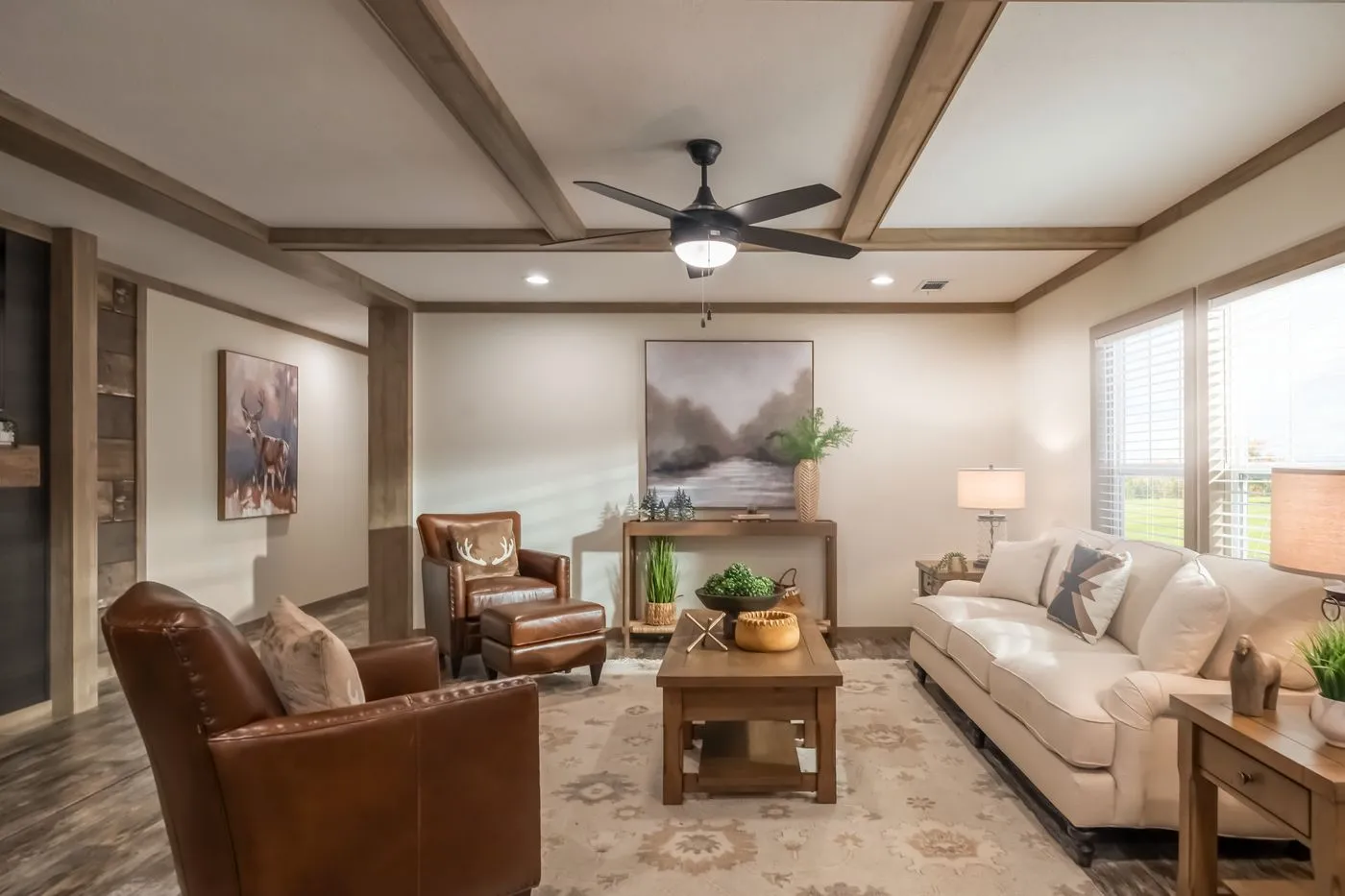 A living room with a white sofa, two brown leather armchairs, a wooden coffee table, potted plants, wall art, and large windows with blinds letting in natural light.