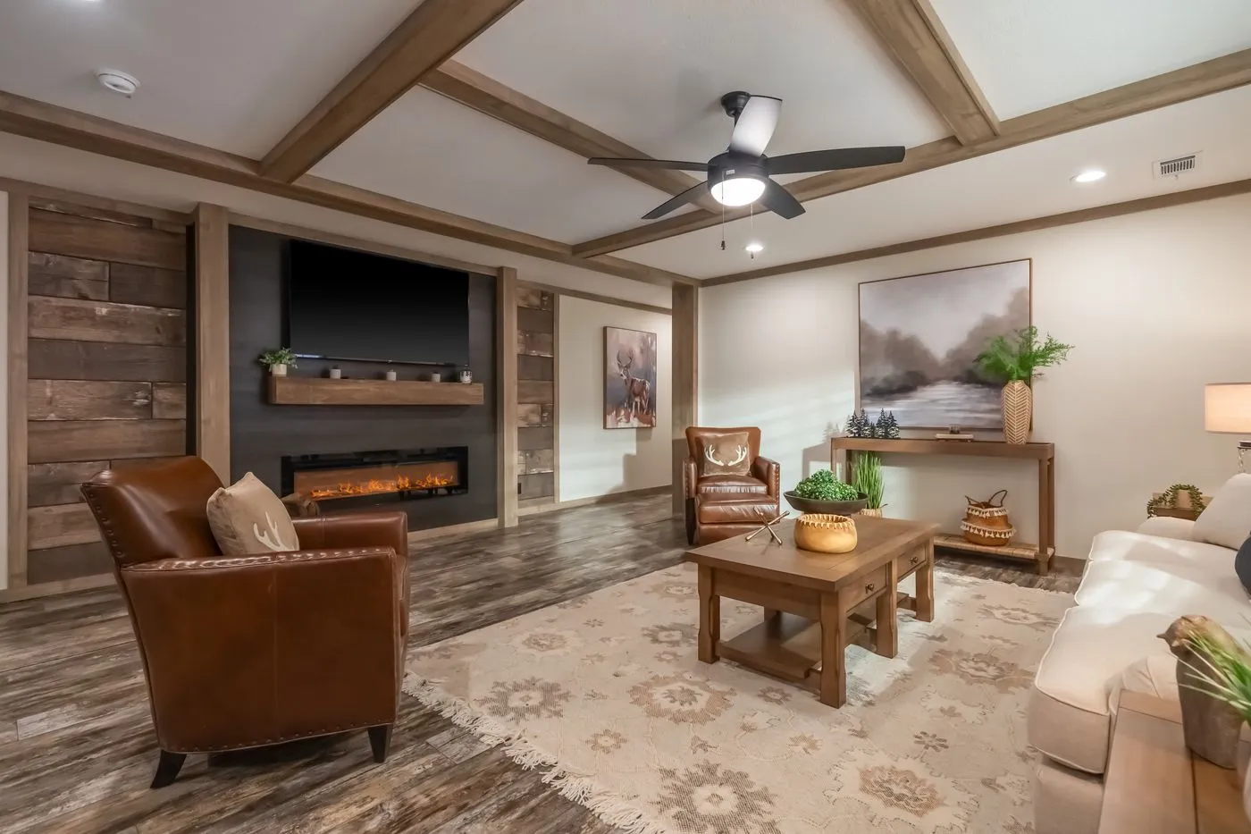 A living room with wooden beams, brown leather chairs, a white sofa, a coffee table, a fireplace with a TV above it, and decor including plants and artwork.