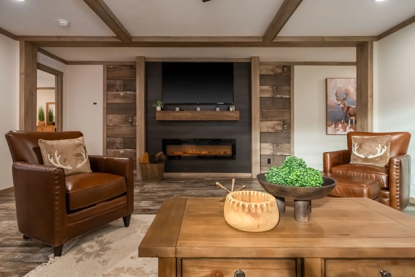 A cozy living room with two brown leather chairs, a wooden coffee table, a wall-mounted TV above a fireplace, and rustic wood accents. Decor includes antler pillows and a bowl of greenery.